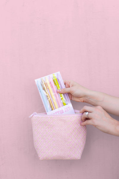 A woman's hands pulling a pack of pens from a pink straw cosmetics pouch.