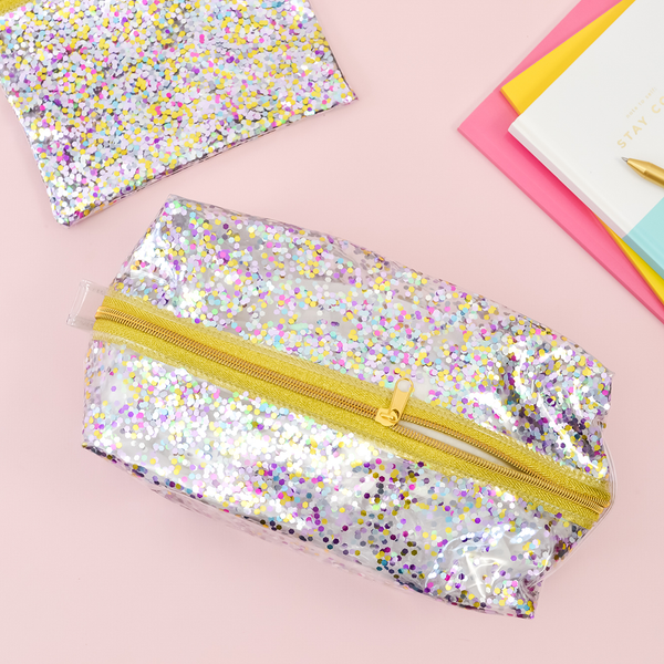 A dual layered clear vinyl confetti dopp kit with a metallic gold zipper halfway zipped. A confetti pouch is in the top left corner and notebooks are seen to the right side. 
