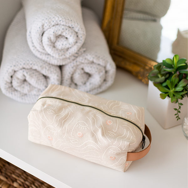 zen ladies doppleganger pouch in tan with a white outline silhouette drawing of a woman's face with blush colored cheeks sitting in a white table with a sage green hairbrush, succulent, white towels and a gold mirror
