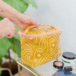 woman with a left arm tattoo closing the zen ladies yellow gold with a white woman's silhouette drawing and blush cheeks makeup tote with handle with makeup sitting on a clear side table next to the bag