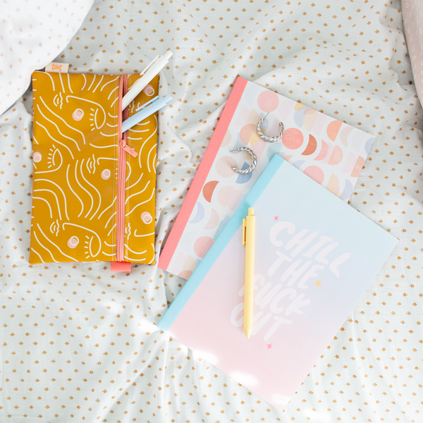 Notebook Set. 1st Notebook Blue to Pink Gradient with Chill The Fuck Out text in white. 2nd Notebook Multi Colored Moon Phase all over pattern. Laid out with a  mustard yellow pouch designed with abstract women faces in white lines and white, banana, and powdered blue colored jotter pens