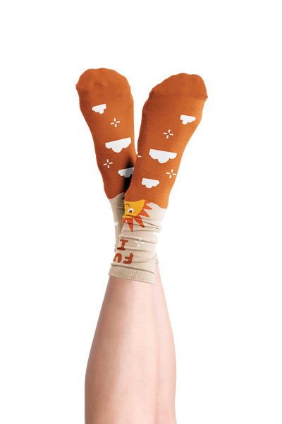 Long socks with a sun rise and the phrase "Fuck it" displayed are the top of the sock. Lower part of the sock is brown with clouds and sparkle stars. Displayed on a person crossing their legs.
