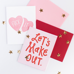 Image of two cards. The one on the left is white with pink hearts and the text "Fuck It". The one on the left is red and white striped with red text "Let's Make Out". Cards are sprinkled with gold star confetti.