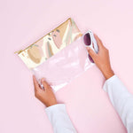 A clear vinyl pouch with the top of the bag having an abstract fruit design in cream colors and muted pastels like green and pink. Paint-like smears are also a part of the design. Pouch is being held by person with sunglasses in front of pink background.