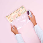 A clear vinyl pouch with the top of the bag having an abstract fruit design in cream colors and muted pastels like green and pink. Paint-like smears are also a part of the design. Pouch is being held by person with sunglasses in front of pink background.
