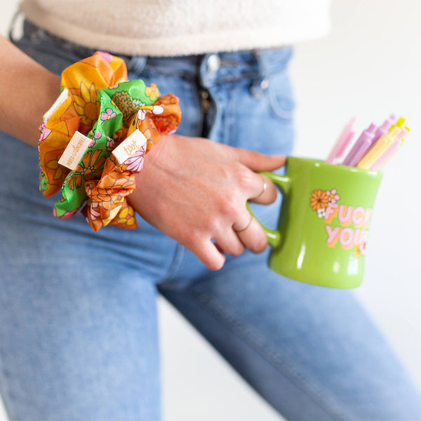 woman holding a fuck you green diner mug with three floral print ripstop scrunchies, one in pink, one in green, and one in brown