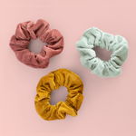 A dusty pink, light blue, and tan scrunchies in corduroy material. All scrunchies are in front of a light pink background.