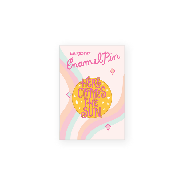 round enamel pin with yellow ground and pink letters saying here comes the sun