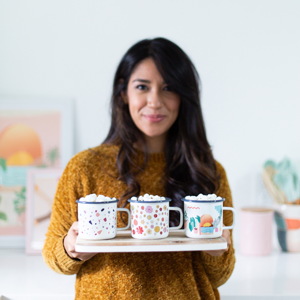 a girl holding three campfire mugs. one with flowers, one with a sunset and plants, and one terrazzo