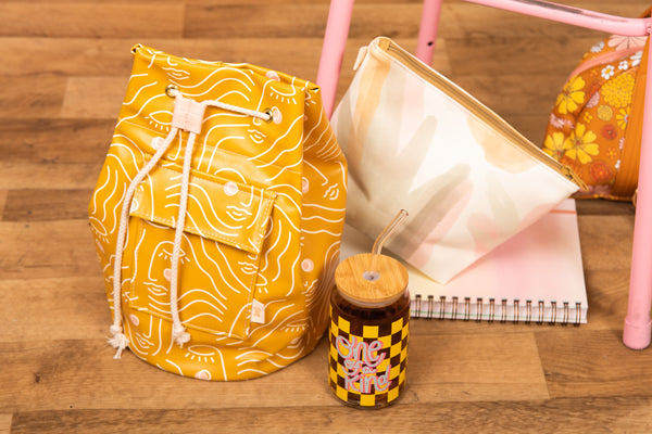 Mustard yellow drawstring bag with white and pale pink face design, next to another glass, pouch, and notebook