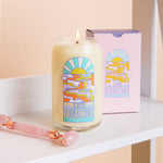 16 oz candle with "Good Vibrations" on it and a melting sunset. It is stood on a shelf next to a rose quartz face roller.