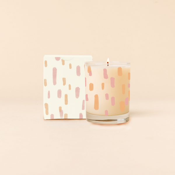 Rock glass candle with muted orange and pink sun drop pattern surrounding candle. Box packaging with same design as candle.  