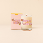 Rock glass candle reading "GREAT VIBES" with colorful cluster flower design at opposite corners of statement. Pastel pink box packaging with same design as candle. 