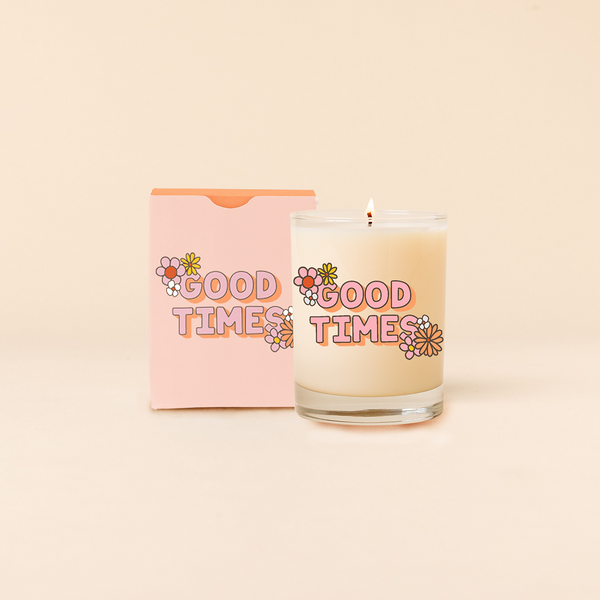 Rock glass candle reading "GOOD TIMES" with colorful cluster flower design at opposite corners of statement. Pastel pink box packaging with same design as candle.