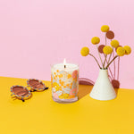 Pastel pink background with rising scallops rock glass candle with misc items on a mustard yellow surface. 
