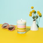 Light blue background with rising scallops rock glass candle with misc items on a mustard yellow surface. 