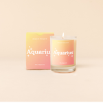 Candle rocks glass with yellow-to-orange ombre decal and text that reads "Aquarius" with minimalist, white sparkle stars surrounding the text; "the unique one" sits at the bottom of the decal. Box packaging with the same design sits behind glass.