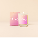Candle rocks glass with cream-to-pink ombre decal and text that reads "Aries" with minimalist, white sparkle stars surrounding the text; "the one who thinks they rule" sits at the bottom of the decal. Box packaging with the same design sits behind glass.