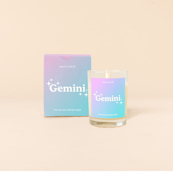Candle rocks glass with lavender-to-bright blue ombre decal and text that reads "Gemini" with minimalist, white sparkle stars surrounding the text; "the one who shines bright" sits at the bottom of the decal. Box packaging with the same design sits behind glass.