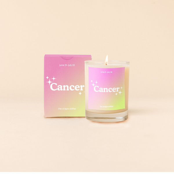Candle rocks glass with bright pink-to-bright green ombre decal and text that reads "Cancer" with minimalist, white sparkle stars surrounding the text; "the shape shifter" sits at the bottom of the decal. Box packaging with the same design sits behind glass.