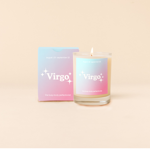 Candle rocks glass with bright blue-to-pink ombre decal and text that reads "Virgo" with minimalist, white sparkle stars surrounding the text; "the busy-body perfectionist" sits at the bottom of the decal. Box packaging with the same design sits behind glass.