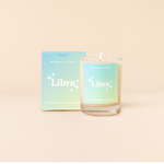 Candle rocks glass with light blue-to-seafoam green ombre decal and text that reads "Libra" with minimalist, white sparkle stars surrounding the text; "the flirtatious, creative, peacemaker" sits at the bottom of the decal. Box packaging with the same design sits behind glass.