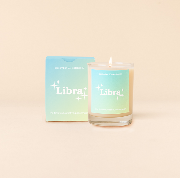 Candle rocks glass with light blue-to-seafoam green ombre decal and text that reads "Libra" with minimalist, white sparkle stars surrounding the text; "the flirtatious, creative, peacemaker" sits at the bottom of the decal. Box packaging with the same design sits behind glass.
