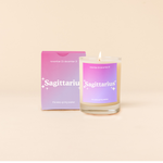 Candle rocks glass with dark pink-to-lavender ombre decal and text that reads "Sagittarius" with minimalist, white sparkle stars surrounding the text; "the easy-going seeker" sits at the bottom of the decal. Box packaging with the same design sits behind glass.