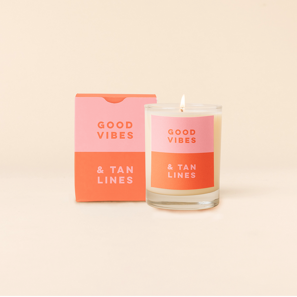 Rocks candle with two tone backdrop. Top half light pink backing with orange text and and bottom half orange backing with pink text reading "GOOD VIBES & TAN LINES". Two tone box packing with same text as candle. 