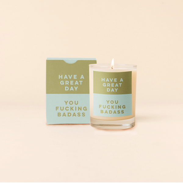Rocks candle with two tone backdrop. Top half green backing with teal text and bottom half teal backing with green text reading "HAVE A GREAT DAY YOU FUCKING BADASS". Two tone box packing with same text as candle.