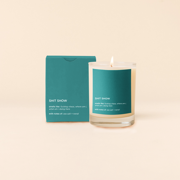 14 oz candle rocks glass with teal decal and text that reads "SHIT SHOW; smells like: fucking chaos, where am i, what am i doing here; with notes of: sea salt + neroli" in white font. Teal box packaging with the same design sits behind the candle.