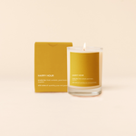 14 oz candle rocks glass with mustard yellowdecal and text that reads "HAPPY HOUR, smells like: fresh cocktails, good music, laughter, with notes of: sparkling yuzu and good times" in white font. Mustard yellow box packaging with the same design sits behind the candle.