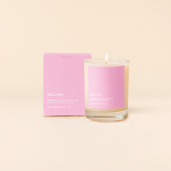 14 oz candle rocks glass with pink decal and text that reads "WELL SHIT, smells like: Hmmmmm that went different in my head, fuck it, with notes of: floral + fig" in white font. Pink box packaging with same design sits behind candle.