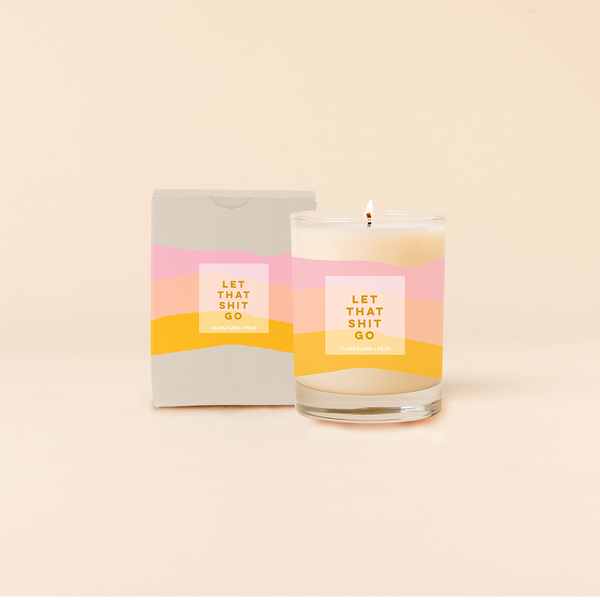 Rocks candle reading "LET THAT SHIT GO" in front of tri-color horizontal stripes around candle. Box packaging with same design and text as candle.