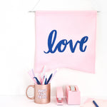 A blush pink canvas hanging wall pennant with "Love" printed on in in navy blue. Displayed with blush pink stationary and a rose gold mug that says "Don't talk to me."