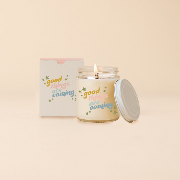 Glass jar candle reading "GOOD THINGS ARE COMING" in multi-color text surrounded by green stars. Box packaging with same design as candle. 