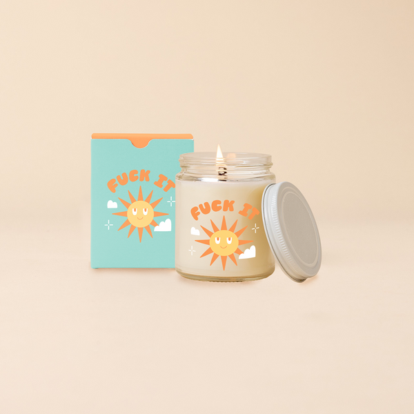 Glass jar candle reading "FUCK IT" in orange bubble letters above sunshine with happy face surrounded by minimal clouds and stars. Blue box packaging with same design as candle. 