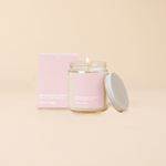 Glass jar candle with lid reading "DEEP BREATHS IN/smells like now let that shit go, peony & amber" white text on light pink background. Light pink box packing with same text as candle. 