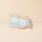 Jar candle with lid; Powder blue, mint, and light blue wavy print wraps around the jar with text that reads "EXHALE THE BS" on the front of the candle. Box packaging with same design sits behind candle.