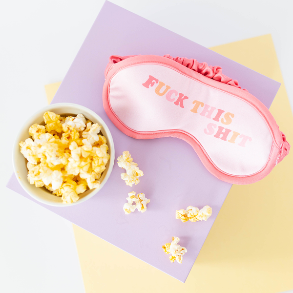 A sleep mask with pink trim that reads "Fuck this shit" in light pink, dark pink and orange text. There is a bowl of popcorn with popcorn pieces scattered around. 