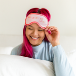 Girl holding a fluffy pillow about to pull her light red and pink sleep mask on that says "Fuck this shit" in pink, red, and orange.