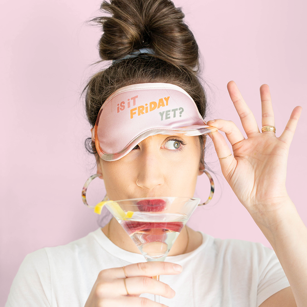 A woman is sipping a martini wearubg a sleep mask that reads "Is it Friday yet" in shades of pink, cream, orange and green.. She is holding it up over one eye so she can see.