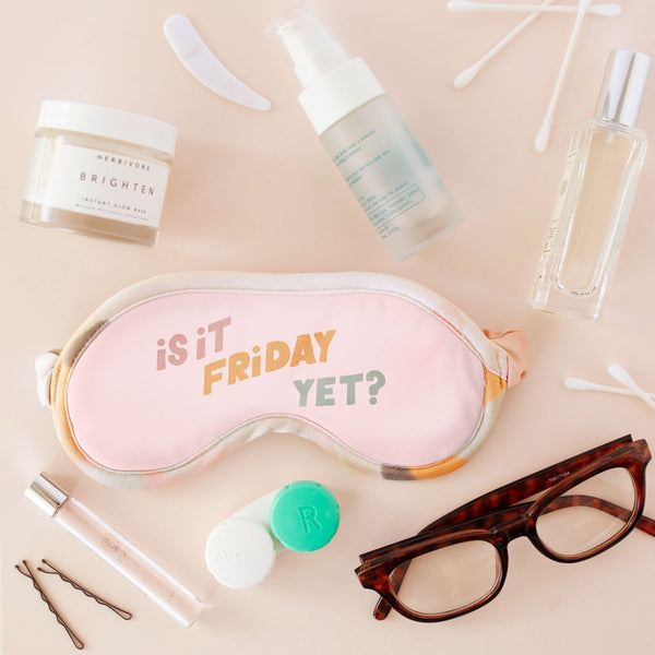 A light pink eye mask with the phrase "Is it Friday yet?" printed on in muted colored letters. Displayed with eye glasses, eye contacts, and cosmetic products.