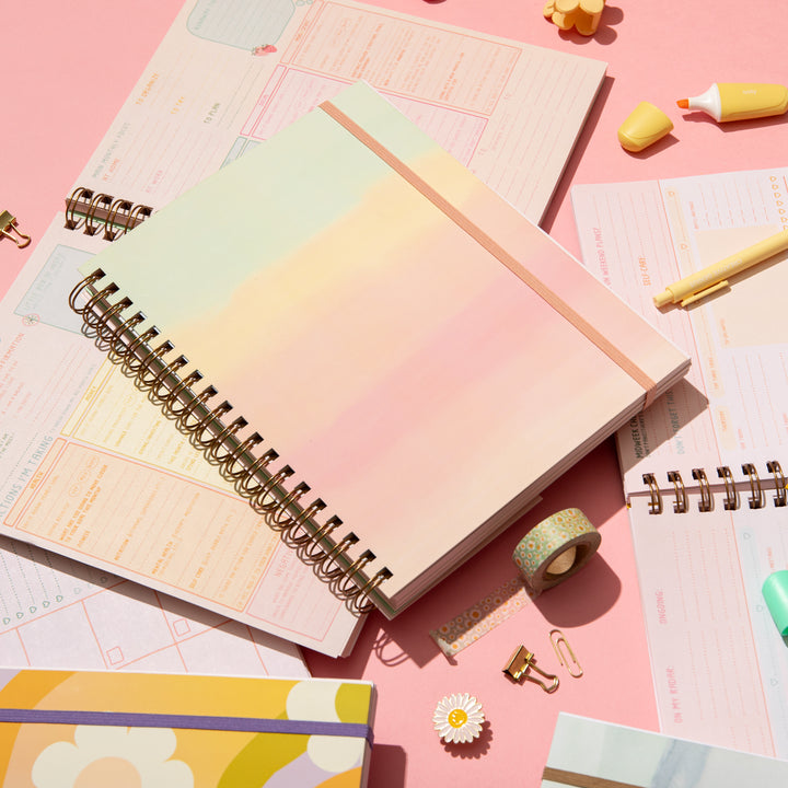 A flat lay on a pink background showcasing a pastel gradient spiral planner with an elastic band, surrounded by open planner pages, yellow and mint highlighters, a yellow pen, washi tape, gold binder clips, and flower-shaped accessories. The scene is bright, colorful, and designed to inspire cheerful planning.