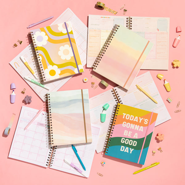 Open and closed undated perpetual planners with stationary essentials surrounding the planners on a pink background.
