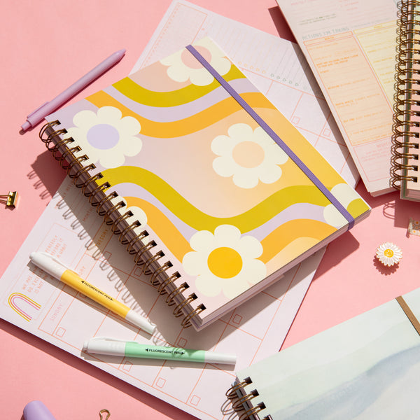 Wavy Daisy Undated Perpetual planner open and closed with pens and highlighters around and a white flower enamel pin on a pink background.