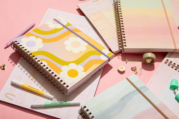 Wavy Daisy, Daybreak, and  Sunset Stripe undated perpetual planner scattered around a pink background closed and open. A white flower enamel pin, flower wash-tape, and highlighters around the planners.