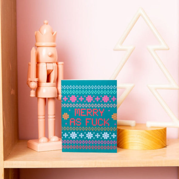 Turquoise "MERRY AS FUCK" in ugly Christmas sweater design, next to a pink nutcracker. 
