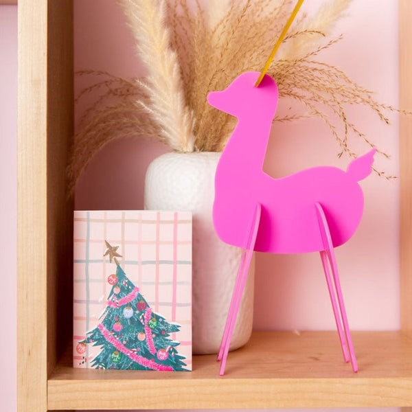 A hot pink reindeer decoration standing next to a slanted Christmas Tree greeting card.