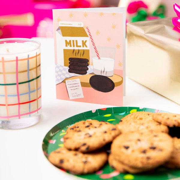 A Milk & Cookies Christmas card next to a plate of cookies and a plaid candle.